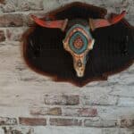 Porte-manteau crâne de vache en résine avec fond en bois et cuir imitation serpent pour décoration esprit motard, avec cornes permettant de déposer casque et gants.