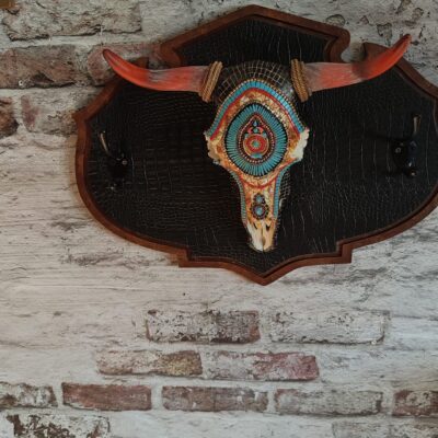 Porte-manteau crâne de vache en résine avec fond en bois et cuir imitation serpent pour décoration esprit motard, avec cornes permettant de déposer casque et gants.